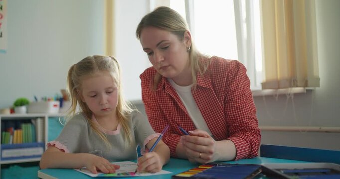 Patient female teacher guiding a young schoolgirl during a writing lesson at a classroom desk, supporting focused learning and creativity