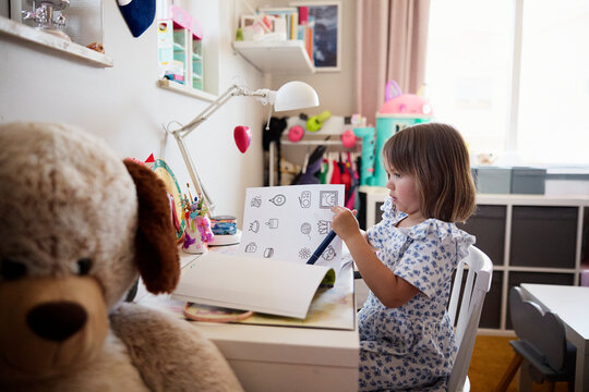 Cute girl sitting with coloring book at study table in bedroom