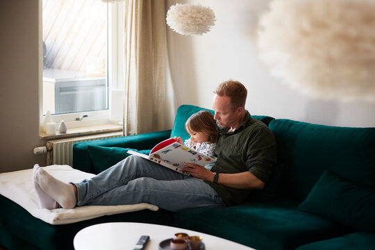 Full length of father reading picture book to daughter while sitting comfortably on sofa in home