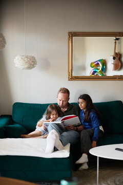 Girls listening to father reading picture book for them while sitting on sofa in living room