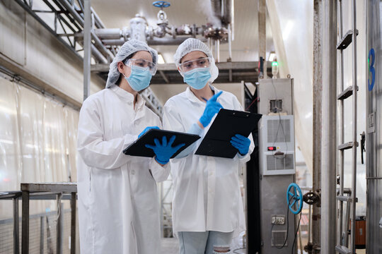 Two female industrial engineers coworkers inspect and examine quality control, production machines in food manufacturing factory, clean and protective uniforms for maintenance, and evaluate operation.