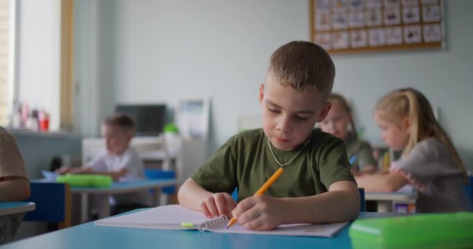 Focused young schoolboy writing with a pencil in a notebook at his classroom desk while classmates study in a bright elementary school setting