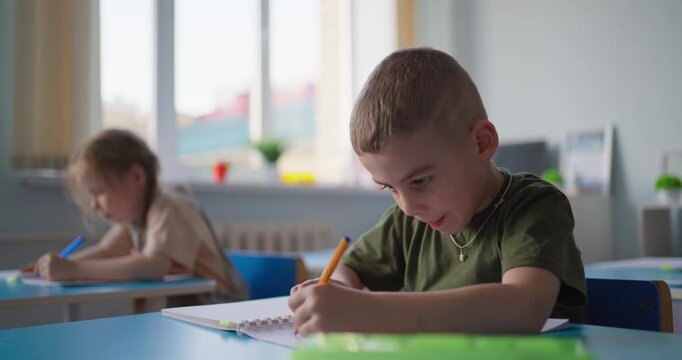 Focused young boy writing with pencil in notebook at classroom desk while classmates study quietly in a bright elementary school setting