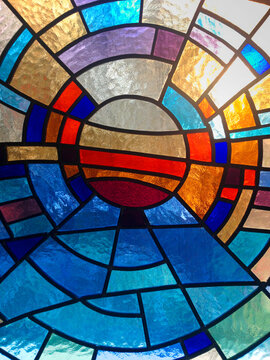 Beautiful stained glass window texture with a circular geometric pattern. Colorful glass mosaic panel backlit by warm sunlight, featuring vibrant blue, red, gold, and orange segments. Abstract art