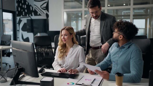 Three joyful multiethnic business people team comparing data stats in documents typing laptop talking happy smiling Caucasian woman businesswoman with Arabian men businessmen talk smile work in office