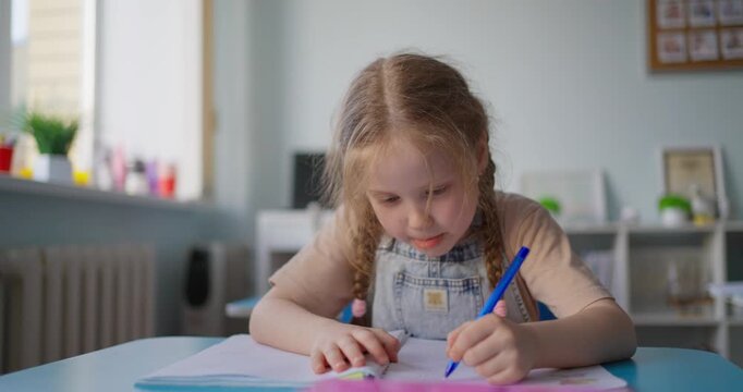Young schoolgirl with braided hair writing in a notebook at a classroom desk while classmates study in a bright early education setting