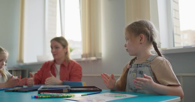 Young girl sitting at a classroom table with crayons and drawings during a creative lesson while a teacher and another child work nearby