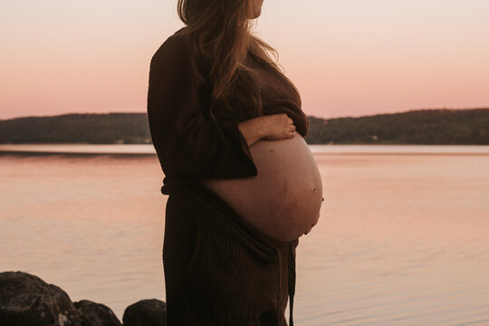 Midsection of pregnant woman touching her belly while standing near river at sunset