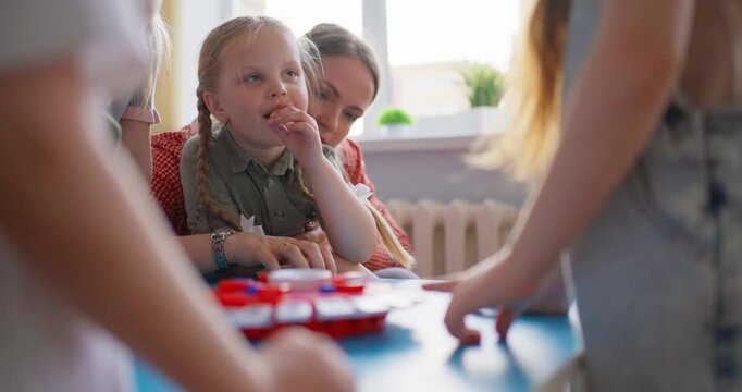 Young children gather around a colorful tabletop game during a supervised classroom activity, learning and playing together in a bright daycare setting