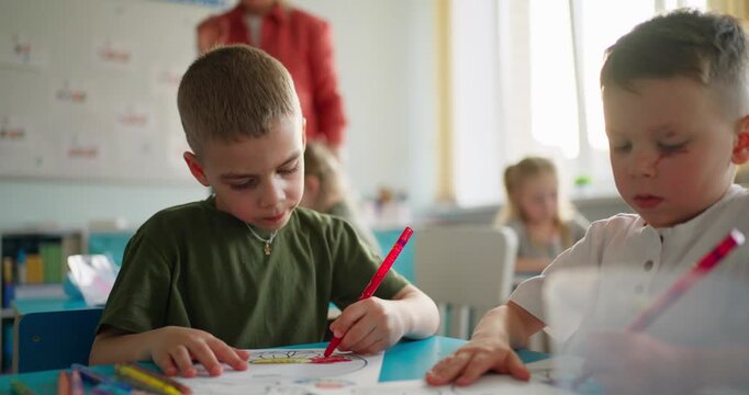 Young schoolchildren drawing with colored pencils at classroom desks during a quiet art lesson, with classmates and teacher softly blurred in background