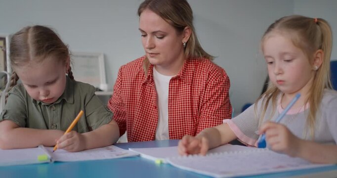Caring teacher supervising two young girls as they practice writing and drawing with pencils at a classroom table during an early learning lesson