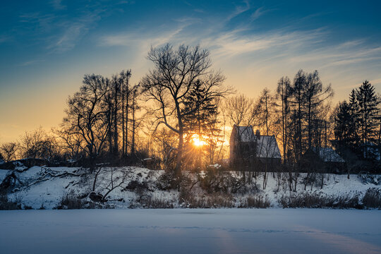 Beautiful winter sunset landscape with sunbeams bursting through bare trees over a small snow-covered house and a frozen lake. Magical golden hour light over winter countryside.