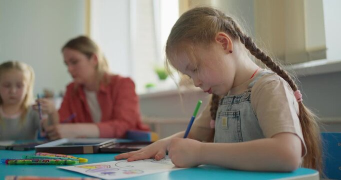 Focused young girl with braided hair coloring a worksheet at a classroom table while classmates and a teacher learn together in the background