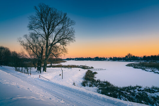 Majestic winter landscape at sunset with a wide frozen river covered in snow. Bare trees standing on the snowy bank under a clear vibrant sky during golden hour.