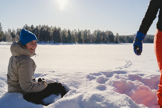 Portrait of happy woman sitting in thick snow on land while enjoying leisure time with friend at sunny day