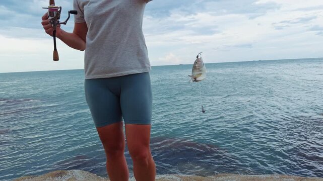 Young Woman Holding Freshly Caught Sea Fish on Fishing Hook Near Tropical Ocean While Standing on Rocky Coastline Under Cloudy Sky, Recreational Fishing Success