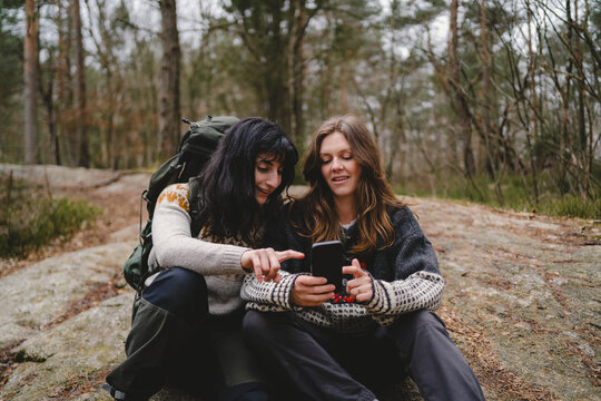 Female friends using smart phone while taking break and sitting on rock in forest