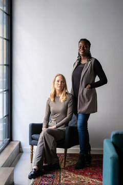 Portrait of smiling female business professional standing near coworker sitting on armchair in office