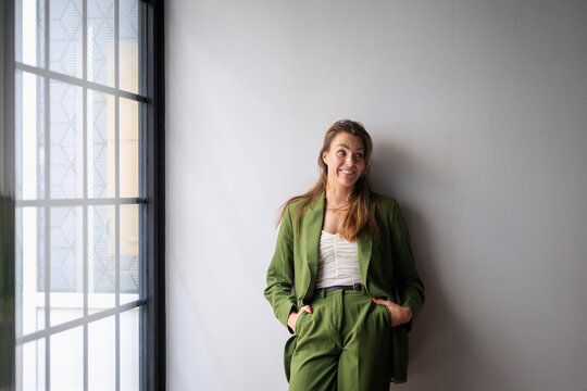 Smiling female business entrepreneur leaning on wall with hands in pocket at office