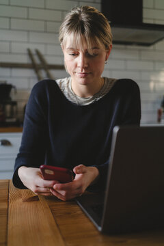 Blond woman using smart phone while sitting with laptop at table in home