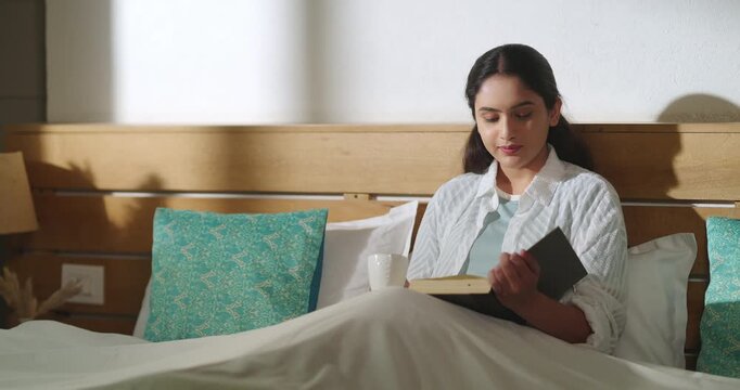 Indian young adult gen z relax calm woman sitting on cozy bed read new novel book story indoor room girl hold cup mug drink hot tea coffee spend time do self care enjoy day take rest break home place