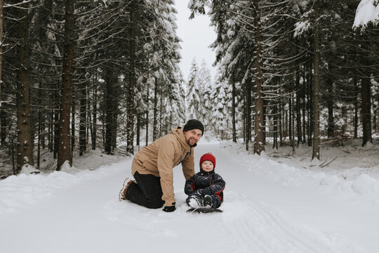 Portrait of smiling man kneeling near toddler son sitting on snow covered road amidst trees