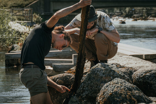 Man examining plant bark with male friend crouching on rocks near lake during camping