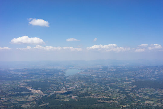High-altitude aerial view of a local pond, surrounding green valleys, and small villages from the top of Mount Ida (Kaz Daglari). Scenic mountain landscape under a bright blue sky with scattered cloud