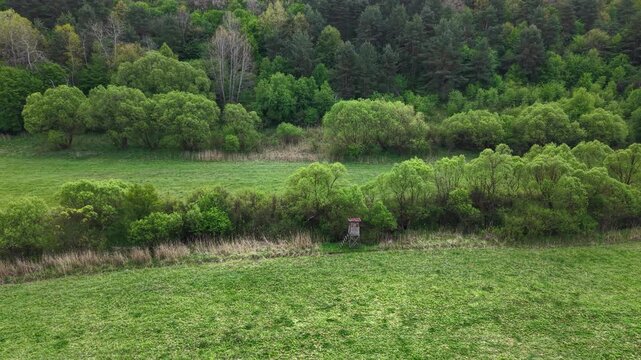 An aerial perspective captures a rustic wooden hunting stand positioned within a lush green meadow, bordered by a dense forest.