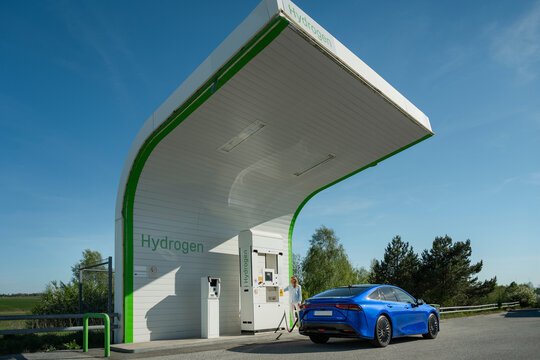 Woman standing near electric car getting charged at hydrogen refueling station on roadside under clear sky