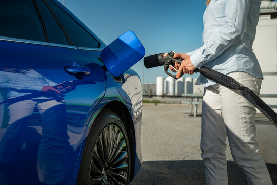 Midsection of woman holding fuel pump while standing near blue electric car at station on sunny day