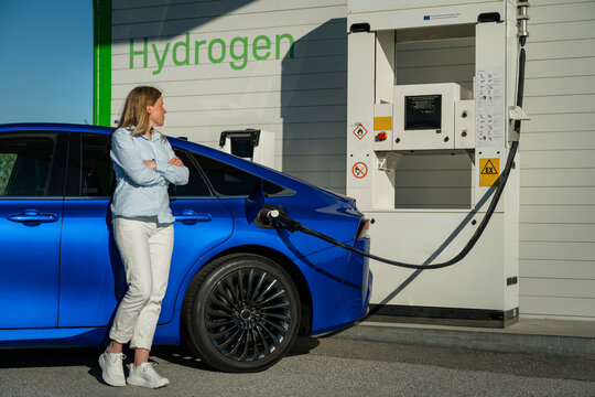 Full length of woman standing with arms crossed while looking at electric car getting charged at station