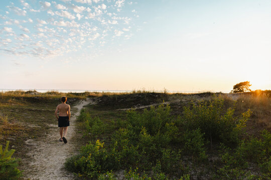 Rear view of shirtless boy walking on footpath amidst grass under cloudy sky at dawn