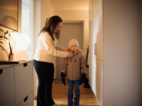Mother zipping jacket of daughter while standing at corridor of house