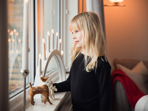 Cute girl with long blond hair standing near needle felted figures kept with electric candle on window sill at home
