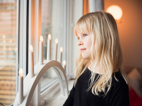 Innocent blond girl looking out through window near electric candles at home