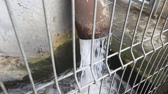Rusty drainage pipe pouring water into an overflowing urban concrete gutter, creating ripples and foam behind a metal security fence, highlighting pollution and waste management issues
