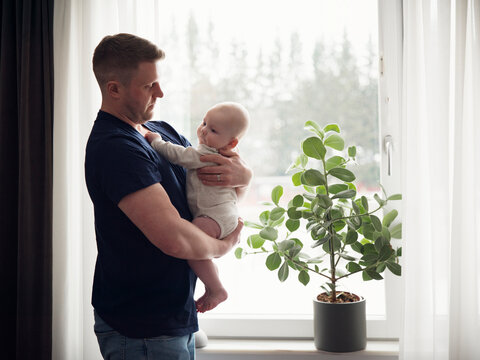 Father spending leisure time with toddler son while standing near window in bedroom at home