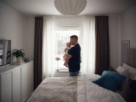 Father kissing baby boy while standing near bed in bedroom at home