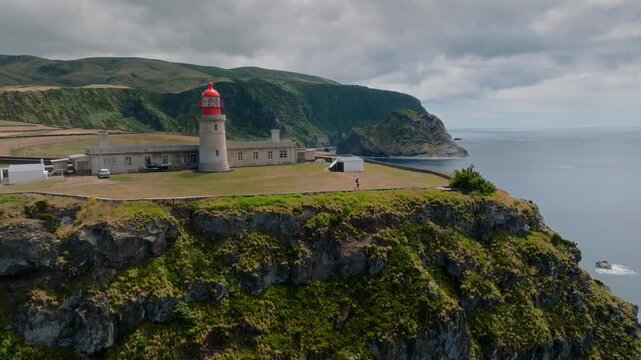 Epic Aerial Drone Flyover of a Solitary Hiker Walking by the Farol de Albarnaz Lighthouse on a Steep Volcanic Cliffside
