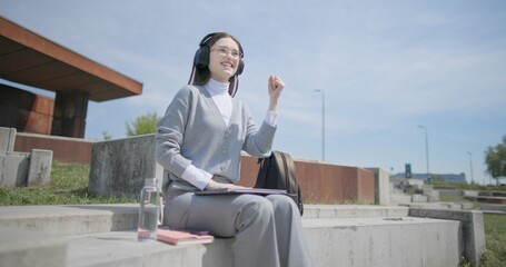 A hand-held shot of a young female student in casual clothes, glasses, and headphones working and typing on a laptop while sitting on a concrete bench in an urban park. © Anatoly Repin
