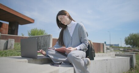 A hand-held shot of a young female student in casual clothes and glasses using a laptop and taking notes with a pen in a notebook in an urban park. © Anatoly Repin
