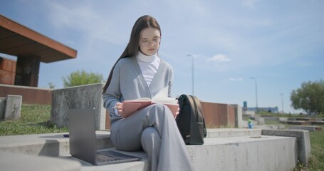 A hand-held portrait of a young female student in casual clothes wearing glasses, sitting on a concrete bench in an urban park, leafing through a notebook and taking notes with a pen. © Anatoly Repin