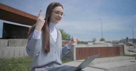 A hand-held shot of a young female student in casual clothes and glasses conducting a video call or conference on a laptop while sitting in an urban park © Anatoly Repin