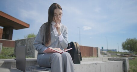 A hand-held portrait of a young female student in casual clothes wearing glasses, sitting on a concrete bench in an urban park, leafing through a notebook and taking notes with a pen. © Anatoly Repin