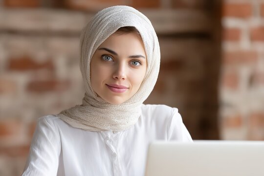 Young professional woman in hijab working on laptop in modern office with brick wall background