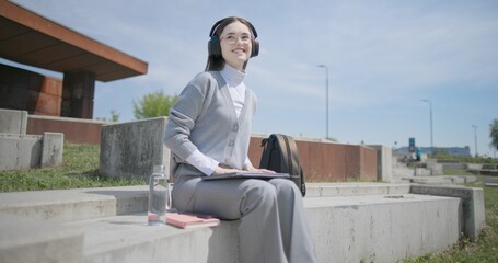 A hand-held shot of a young female student in casual clothes, glasses, and headphones working and typing on a laptop while sitting on a concrete bench in an urban park. © Anatoly Repin