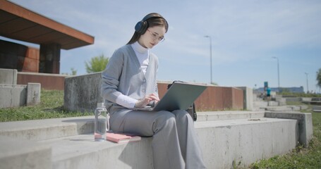 A hand-held shot of a young female student in casual clothes, glasses, and headphones working and typing on a laptop while sitting on a concrete bench in an urban park. © Anatoly Repin