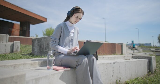 A hand-held shot of a young female student in casual clothes, glasses, and headphones working and typing on a laptop while sitting on a concrete bench in an urban park.