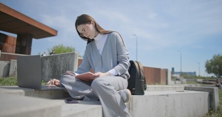A hand-held shot of a young female student in casual clothes and glasses using a laptop and taking notes with a pen in a notebook in an urban park. © Anatoly Repin